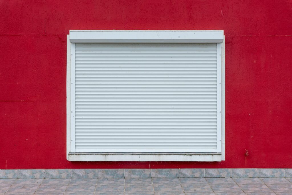 A minimalist exterior view of a white roller shutter on a vibrant red wall.