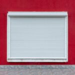 A minimalist exterior view of a white roller shutter on a vibrant red wall.